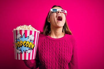 Obraz premium Young beautiful girl watching movie using 3d glasses eating box with popcorns angry and mad screaming frustrated and furious, shouting with anger. Rage and aggressive concept.