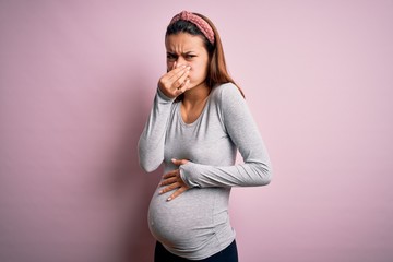 Young beautiful teenager girl pregnant expecting baby over isolated pink background smelling something stinky and disgusting, intolerable smell, holding breath with fingers on nose. Bad smell