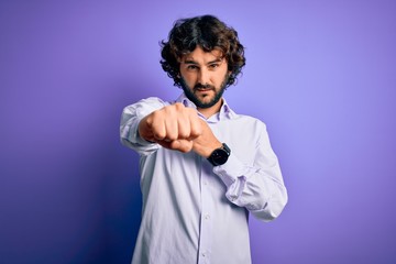 Young handsome business man with beard wearing shirt standing over purple background Punching fist to fight, aggressive and angry attack, threat and violence
