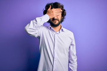 Young handsome business man with beard wearing shirt standing over purple background covering eyes with hand, looking serious and sad. Sightless, hiding and rejection concept
