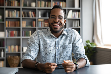 Headshot portrait of happy african American man in glasses sit at desk have video call on gadget,...