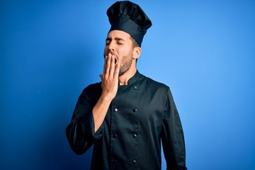 Young handsome chef man with beard wearing cooker uniform and hat over blue background bored yawning tired covering mouth with hand. Restless and sleepiness.