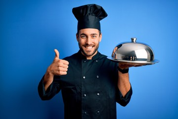 Young cooker man with beard wearing uniform holding tray with dome over blue background happy with big smile doing ok sign, thumb up with fingers, excellent sign