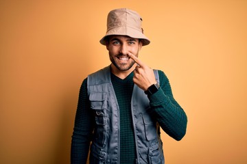 Handsome tourist man with beard on vacation wearing explorer hat over yellow background Pointing with hand finger to face and nose, smiling cheerful. Beauty concept