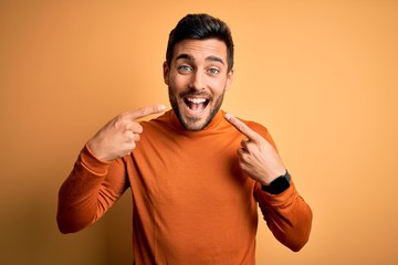 Young handsome man with beard wearing casual sweater standing over yellow background smiling cheerful showing and pointing with fingers teeth and mouth. Dental health concept.