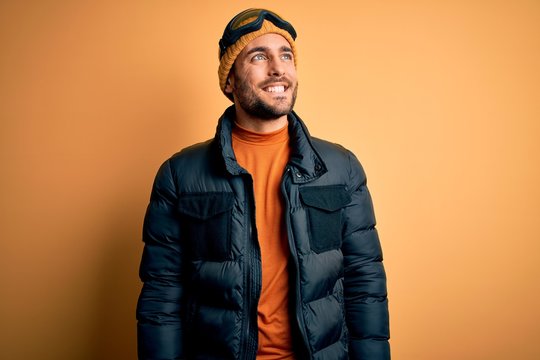 Young handsome skier man with beard wearing snow sportswear and ski goggles looking away to side with smile on face, natural expression. Laughing confident.