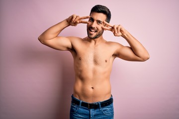 Fototapeta premium Young handsome strong man with beard shirtless standing over isolated pink background Doing peace symbol with fingers over face, smiling cheerful showing victory