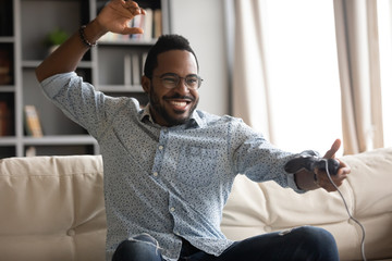 Happy African American young man in glasses sit on couch at home play video game, overjoyed...