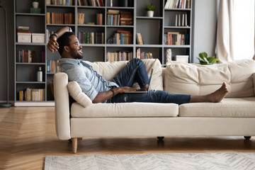 Smiling young African American man relax on comfortable couch in living room using modern laptop,...