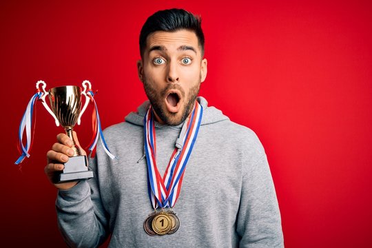 Young handsome succesful man holding trophy wearing medals over red background scared in shock with a surprise face, afraid and excited with fear expression