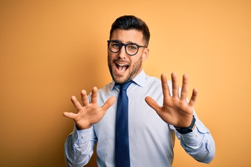 Young handsome businessman wearing tie and glasses standing over yellow background afraid and terrified with fear expression stop gesture with hands, shouting in shock. Panic concept.