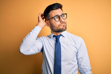 Young handsome businessman wearing tie and glasses standing over yellow background confuse and...