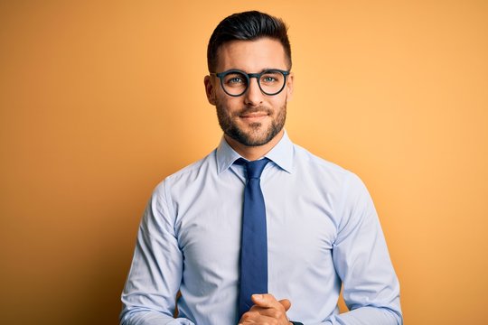 Young handsome businessman wearing tie and glasses standing over yellow background with hands together and crossed fingers smiling relaxed and cheerful. Success and optimistic - Powered by Adobe