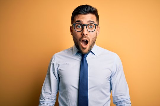 Young handsome businessman wearing tie and glasses standing over yellow background afraid and shocked with surprise and amazed expression, fear and excited face.