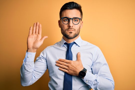 Young handsome businessman wearing tie and glasses standing over yellow background Swearing with hand on chest and open palm, making a loyalty promise oath