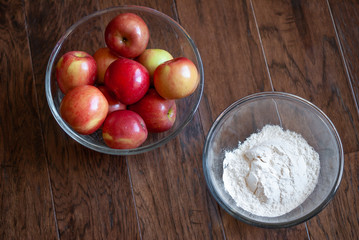 Clear glass bowl of apples and clear glass bowl of flour positioned on a dark wood background