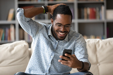 Happy african American man sit on couch at home read good news online on smartphone gadget, smiling biracial male in glasses feel euphoric get pleasant unexpected text or message on cellphone - obrazy, fototapety, plakaty