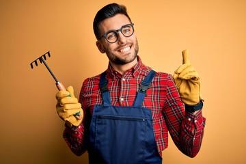 Young gardener man wearing working apron using gloves and tool over yellow background surprised with an idea or question pointing finger with happy face, number one