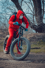 Fototapeta na wymiar Young man in medical mask and gloves, ridding on bicycle in countryside. Male in red suit protecting yourself from diseases on walk. Concept of threat of coronavirus epidemic infection.