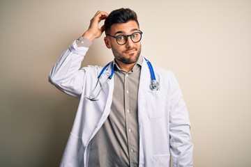 Fototapeta premium Young doctor man wearing glasses, medical white robe and stethoscope over isolated background confuse and wonder about question. Uncertain with doubt, thinking with hand on head. Pensive concept.