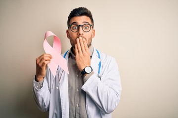 Young doctor man wearing stethoscope holding pink ribbon about cancer over isolated background cover mouth with hand shocked with shame for mistake, expression of fear, scared in silence