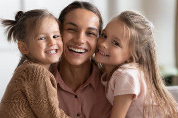 Head shot close up young mixed race woman nanny babysitter touching cheeks with cute little children kids, looking at camera. Portrait of young mother enjoying sweet tender moment with daughters.