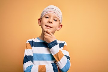 Young little caucasian kid injured wearing medical bandage on head over yellow background looking confident at the camera with smile with crossed arms and hand raised on chin. Thinking positive.