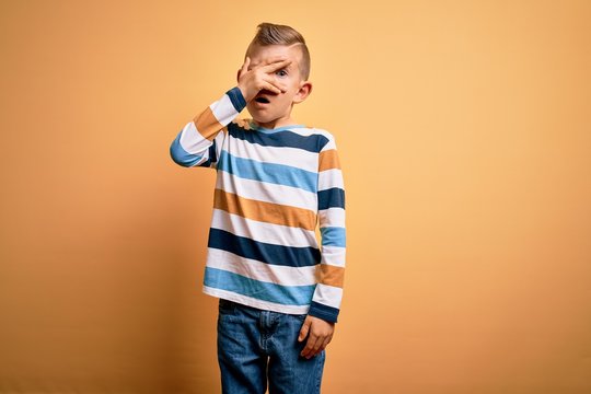 Young little caucasian kid with blue eyes wearing colorful striped shirt over yellow background peeking in shock covering face and eyes with hand, looking through fingers with embarrassed expression.