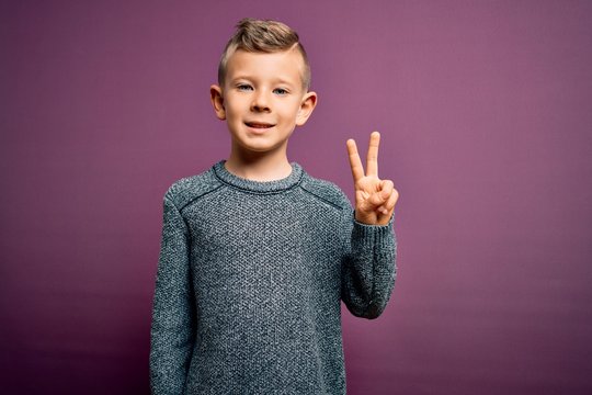 Young little caucasian kid with blue eyes wearing winter sweater standing over purple background smiling with happy face winking at the camera doing victory sign. Number two.