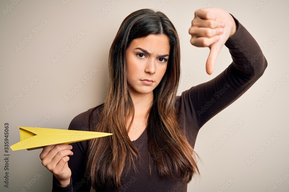 Poster Young beautiful girl holding paper plane standing over isolated white background with angry face, negative sign showing dislike with thumbs down, rejection concept