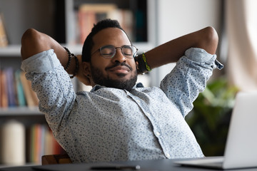 Happy african American male employee lean in chair at workplace relax daydreaming or taking nap,...