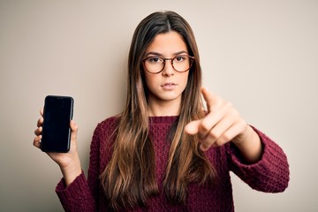 Young beautiful girl wearing glasses showing smartphone over isolated white background pointing...