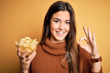 Young beautiful girl holding bowl with chips potatoes standing over yellow background doing ok sign with fingers, excellent symbol