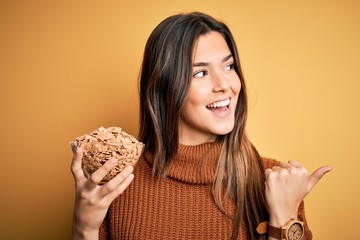 Young beautiful girl holding bowl of healthy cornflakes cereals over yellow background pointing and showing with thumb up to the side with happy face smiling