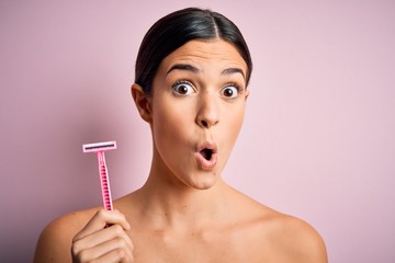 Young beautiful girl using shaver for depilation standing over isolated pink background scared in...
