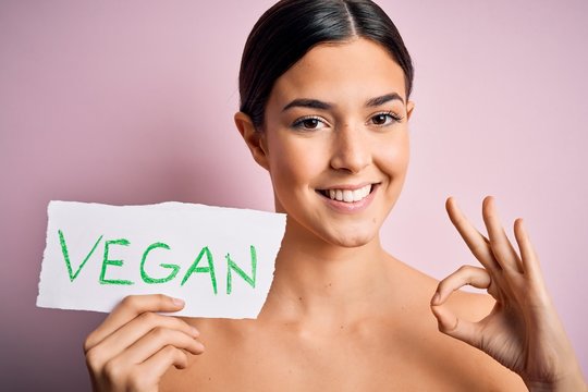 Young beautiful girl holding paper with vegan message over isolated pink background doing ok sign with fingers, excellent symbol