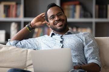 Portrait of happy biracial young man in glasses sit rest on couch at home working on laptop,...