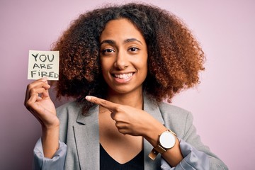 Young african american woman with afro hair holding paper with you are fired message very happy pointing with hand and finger