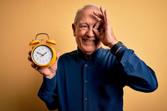 Senior grey haired man holding vintage alarm clock over yellow background with happy face smiling doing ok sign with hand on eye looking through fingers