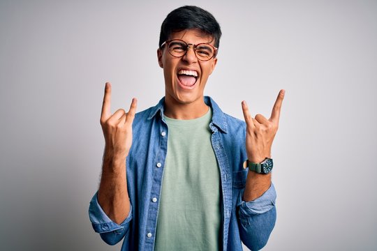 Young handsome man wearing casual shirt and glasses over isolated white background shouting with crazy expression doing rock symbol with hands up. Music star. Heavy concept.