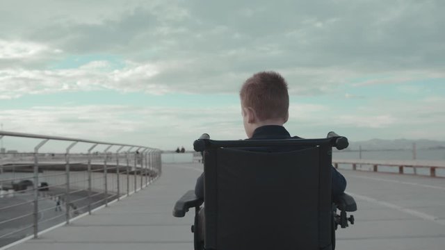 Back middle shot of disable kid in electric wheelchair in the walk. Handicapped boy outdoor