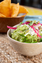 A closeup view of a cup of seasoned guacamole, with a bowl of chips in the background, in a restaurant or kitchen setting.