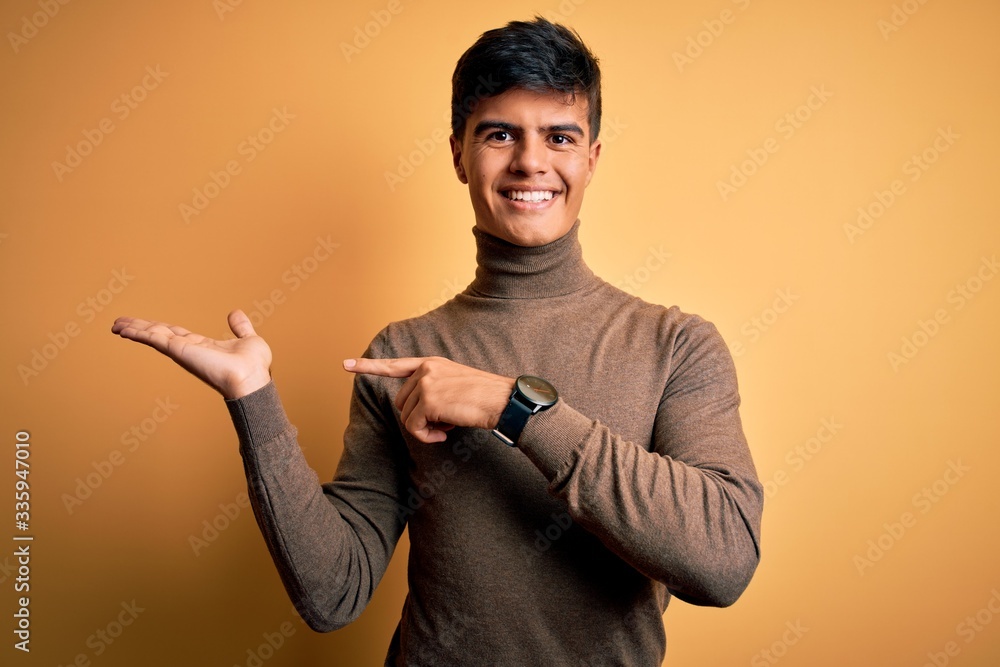 Canvas Prints Young handsome man wearing casual turtleneck sweater over isolated yellow background amazed and smiling to the camera while presenting with hand and pointing with finger.