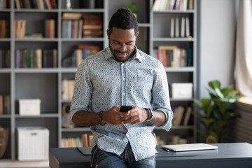 Smiling african American young man stand at desk surfing browsing wireless internet on smartphone...