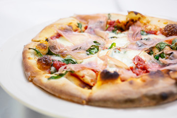 A closeup view of a pizza pie with prosciutto toppings, in a restaurant or kitchen setting.