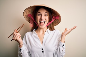 Young beautiful redhead woman wearing asian traditional hat holding wooden chopsticks very happy and excited, winner expression celebrating victory screaming with big smile and raised hands