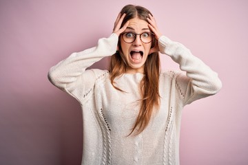 Young beautiful redhead woman wearing casual sweater and glasses over pink background Crazy and scared with hands on head, afraid and surprised of shock with open mouth