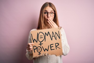 Young beautiful redhead woman asking for women rights holding banner over pink background cover mouth with hand shocked with shame for mistake, expression of fear, scared in silence, secret concept