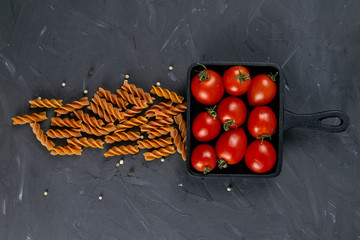 Raw Italian pasta fusilli with cherry tomatoes in frying pan on dark background from above. Flat lay