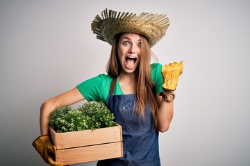 Young beautiful redhead farmer woman wearing apron and hat holding box with plants screaming proud...